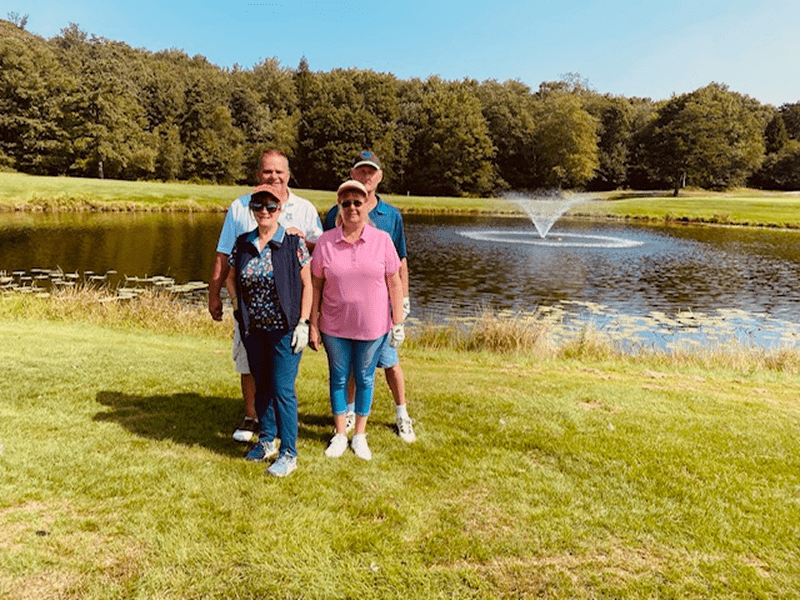 Four women standing on grass near a pond with a fountain on a sunny day.