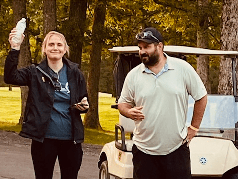 Two people standing outdoors near a golf cart, smiling and holding drinks.