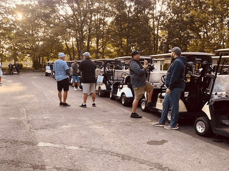 Group of men standing near golf carts on a sunlit path.