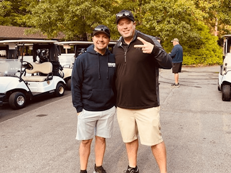 Two men smiling outdoors near golf carts.