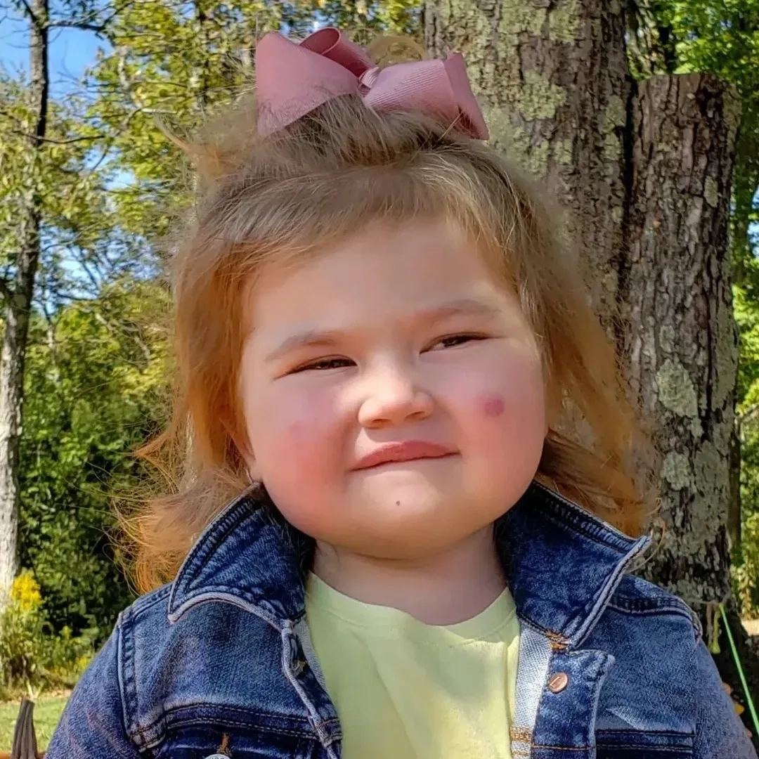 Smiling toddler girl with a pink bow and denim jacket outdoors.